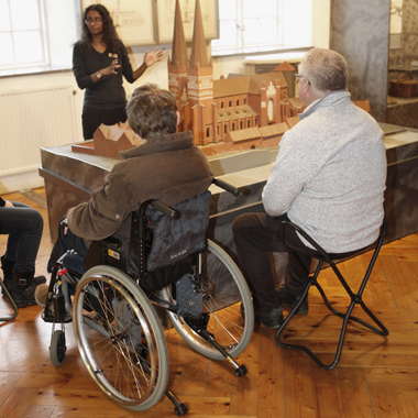 Två män lyssnar på en kvinna som berättar i en utställning.  Den ena sitter i en rullstol , den andre på en pall.