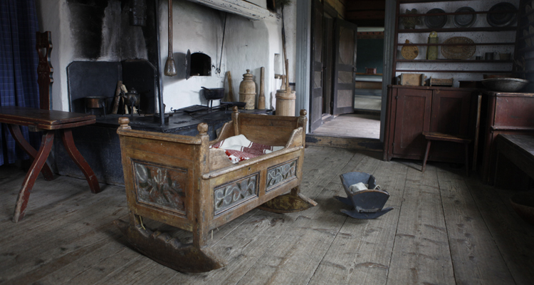 Interiör från 1800-talet, i bakgrunden en öppen spis och en tallriskhylla, i förgrunden en vagga och en leksaksvagga.
